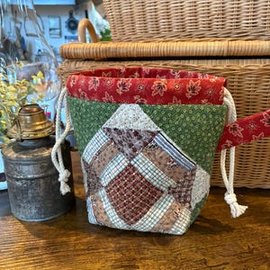 Può includere: Una borsa con coulisse in tessuto patchwork con interno a fantasia floreale rosso ed esterno verde. La borsa presenta un design geometrico sul davanti con vari quadrati di tessuto nei toni del marrone, bianco e blu. La corda bianca viene utilizzata per il cordino.