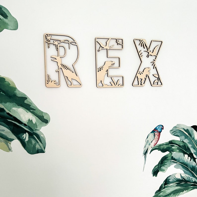 May include: Wooden letters spelling "REX" with dinosaur cutouts. The letters are light brown and have a natural wood finish. The dinosaurs are cut out of the letters and are facing different directions. The letters are on a white background.