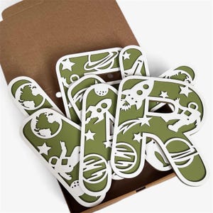 May include: A collection of green and white space-themed letter cutouts. Each letter displays a different space-related graphic, including planets, rockets, astronauts, and stars. The letters are inside a brown cardboard box.