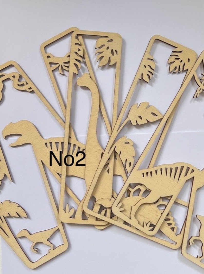 May include: A set of six wooden bookmarks featuring dinosaur silhouettes and tropical leaves. The bookmarks are laser cut and have a natural wood finish. The number "No2" is visible on one of the bookmarks.
