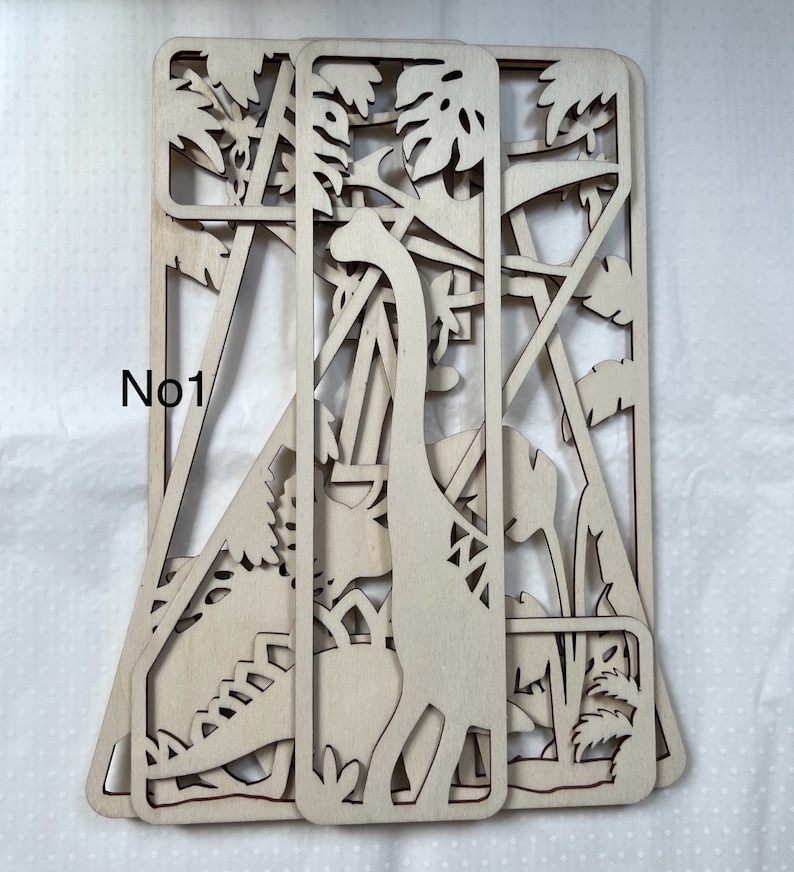 May include: A set of three wooden laser cut panels featuring a dinosaur and jungle foliage. The panels are layered to create a 3D effect. The text "No1" is visible on the left side of the image.