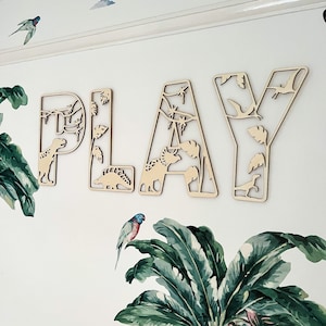 May include: Wooden wall decor spelling out the word "PLAY" with a dinosaur and jungle theme. The letters are cut out and have a natural wood finish.