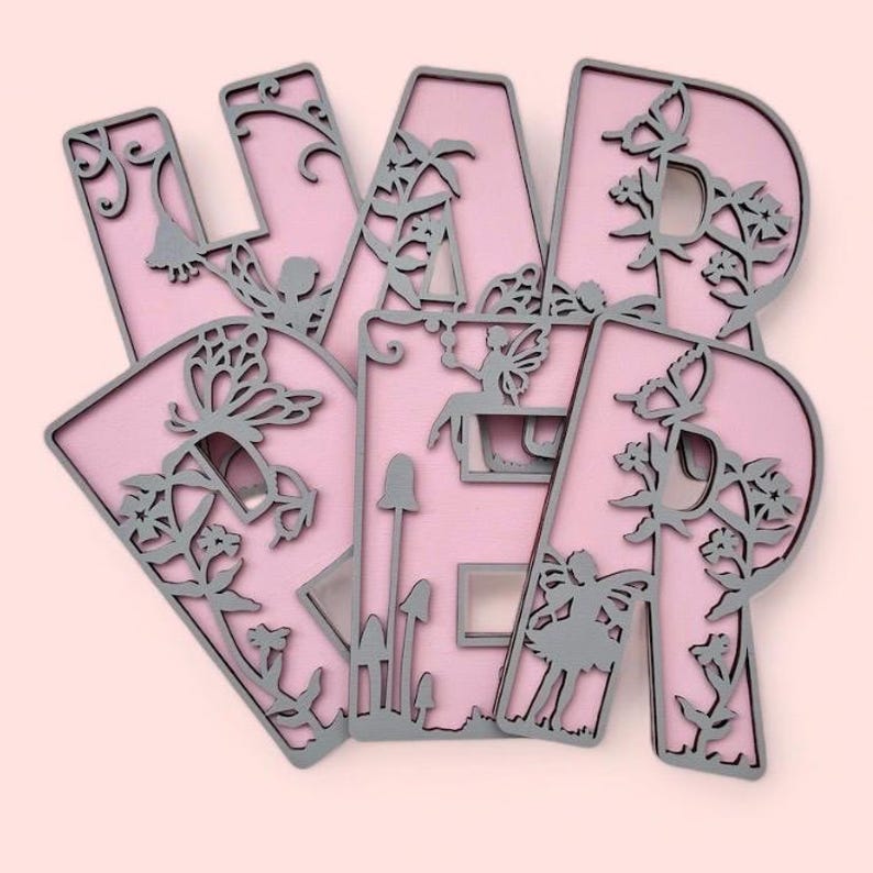 May include: Pink wooden letters spelling "F A I R Y" with a fairy and butterfly cutouts. The letters have a gray outline and are decorated with floral designs.