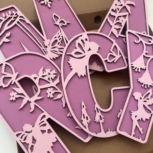 May include: Pink wooden letters with a fairy and floral design. The letters are cut out and have a delicate, intricate design.
