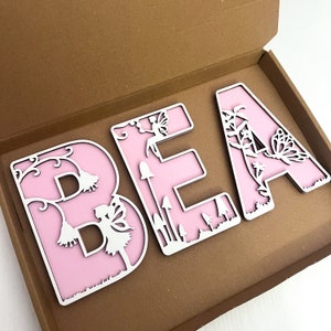 May include: Three pink wooden letters spelling "BEA" with white cutout designs of fairies, butterflies, flowers, and mushrooms.