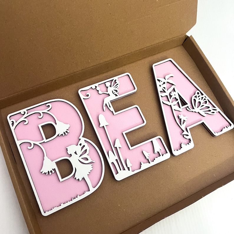 May include: Three pink wooden letters spelling "BEA" with white cutout designs of fairies, butterflies, flowers, and mushrooms.