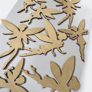 May include: A collection of wooden fairy cutouts in various poses. The fairies have detailed wings and are made of light-colored wood. The cutouts are scattered across a white surface, showcasing their delicate design and whimsical theme.