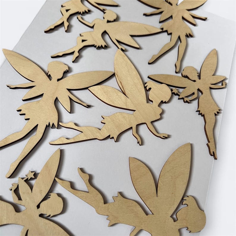 May include: A collection of wooden fairy cutouts in various poses. The fairies have detailed wings and are made of light-colored wood. The cutouts are scattered across a white surface, showcasing their delicate design and whimsical theme.