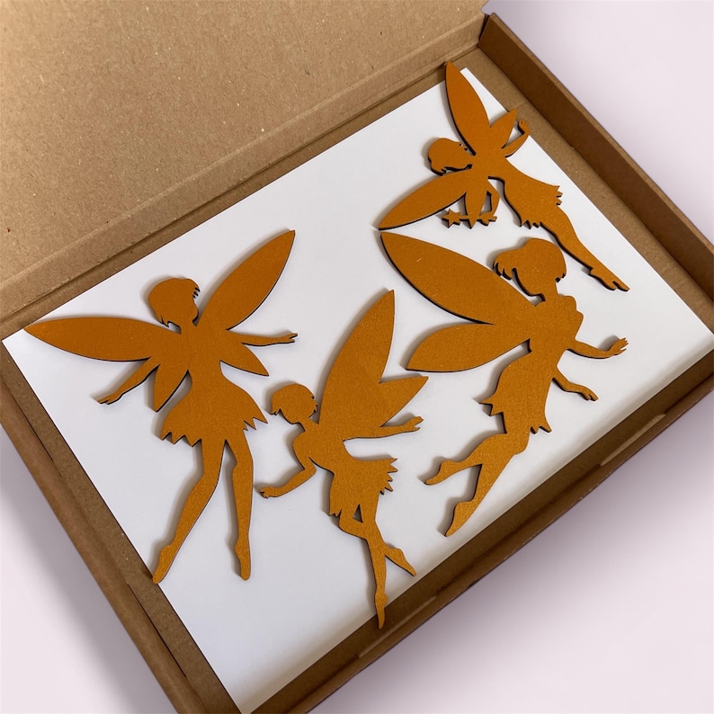 May include: Four wooden fairy cutouts in a cardboard box. The fairies are a warm brown color, with detailed wings and flowing dresses. They are arranged on a white surface inside the box, ready for use in crafts or decor.