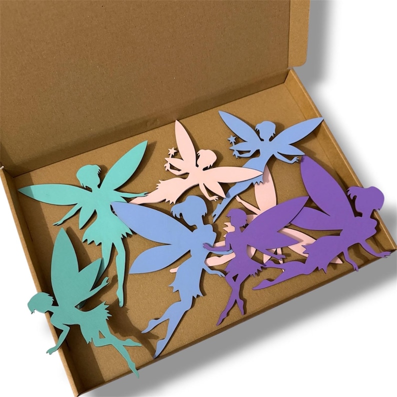 May include: A collection of colorful fairy cutouts in a cardboard box. The fairies are in shades of teal, light blue, pink, and purple. Each fairy has detailed wings and a silhouette design. The box is open, revealing the fairies inside.