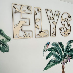 May include: Wooden letters spelling the name "ELYS" decorated with dinosaur and tropical leaf cutouts.