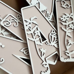 May include: A set of five wooden letters with a delicate floral and butterfly design. The letters are painted a light pink color and have a white cutout design. The letters are arranged in a random order.