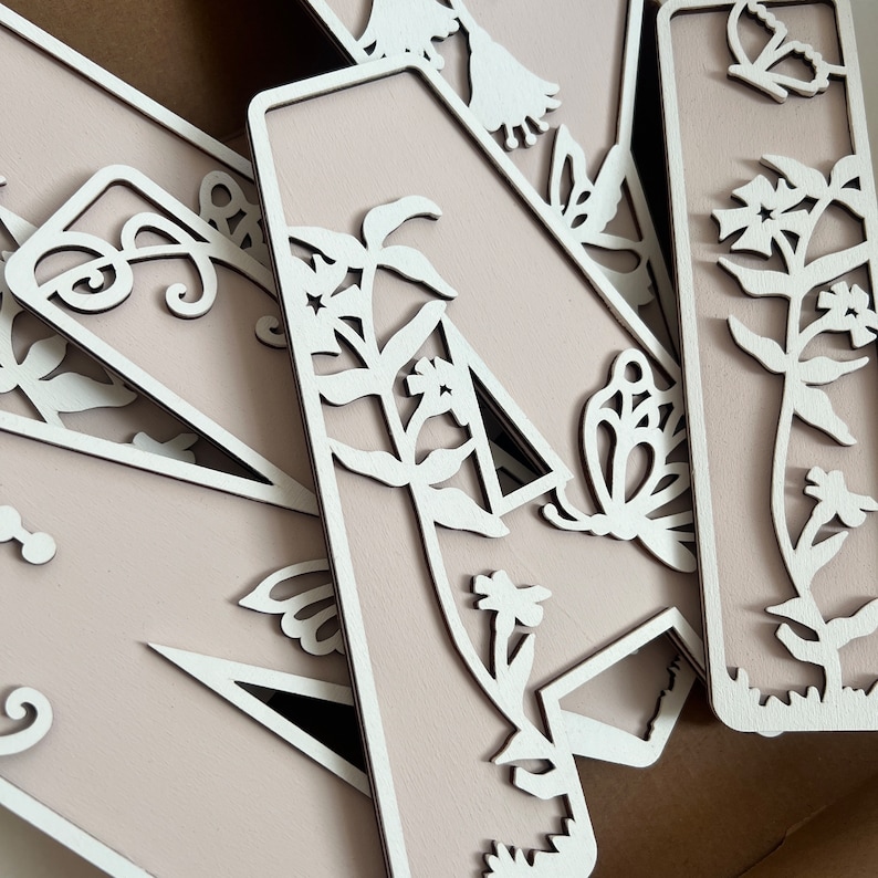May include: A set of five wooden letters with a delicate floral and butterfly design. The letters are painted a light pink color and have a white cutout design. The letters are arranged in a random order.