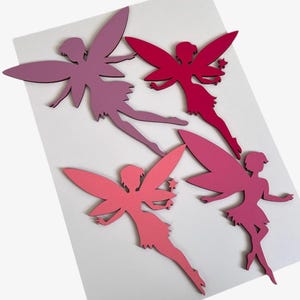 May include: Four wooden fairy cutouts in shades of pink and purple. Each fairy has large wings and a flowing skirt. One fairy holds a star-shaped wand. The fairies are arranged on a white background.