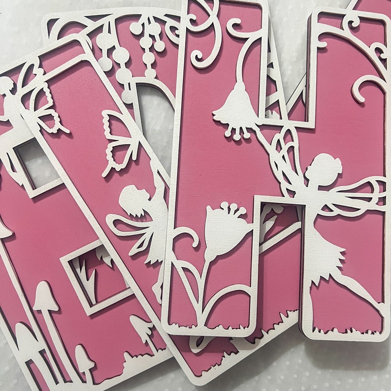 May include: Pink wooden letters with a fairy and flower design. The letters are cut out with a delicate design and are perfect for a child's room.