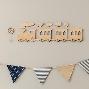 May include: Wooden train wall decor with a train engine, train cars, and cloud cutouts. A railroad crossing sign is to the left. Below, a garland of blue and tan pennant flags hangs from a string.