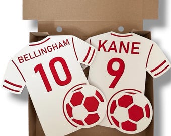 May include: Two white and red flat wooden cutouts shaped like football shirts inside a brown cardboard box. One shirt features the name BELLINGHAM and the number 10, while the other displays the name KANE and the number 9. Each shirt includes a football.
