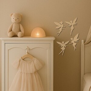 May include: A beige nursery scene features a teddy bear, a glowing dome-shaped lamp, and a white dresser. Four fairy wall decorations are affixed to the wall. A light pink dress hangs on a wooden hanger, adding a touch of whimsy to the decor.