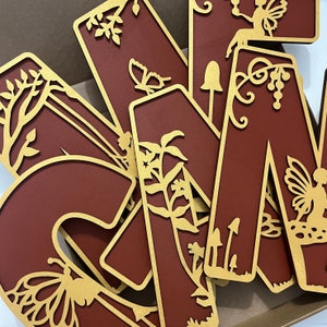 May include: A set of wooden letters in a burgundy color with gold accents. The letters are decorated with intricate cut-out designs of butterflies, flowers, and vines. The letters are arranged in a random order.