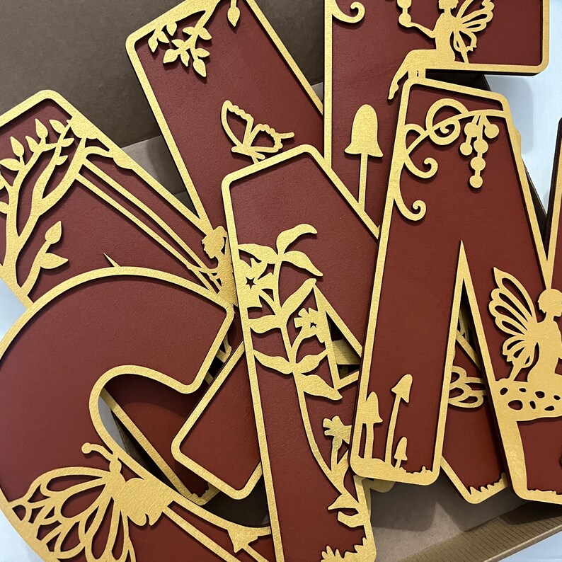 May include: A set of wooden letters in a burgundy color with gold accents. The letters are decorated with intricate cut-out designs of butterflies, flowers, and vines. The letters are arranged in a random order.
