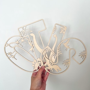 May include: Wooden cutout letters spelling "V", "I", and "V" with dinosaur and plant designs. The letters are layered on top of each other.
