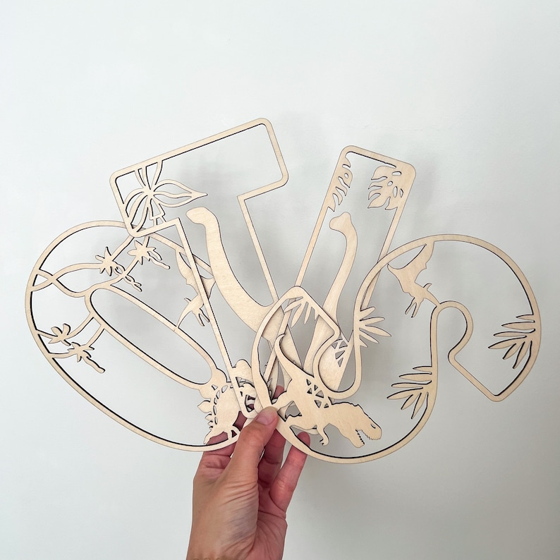 May include: Wooden cutout letters spelling "V", "I", and "V" with dinosaur and plant designs. The letters are layered on top of each other.
