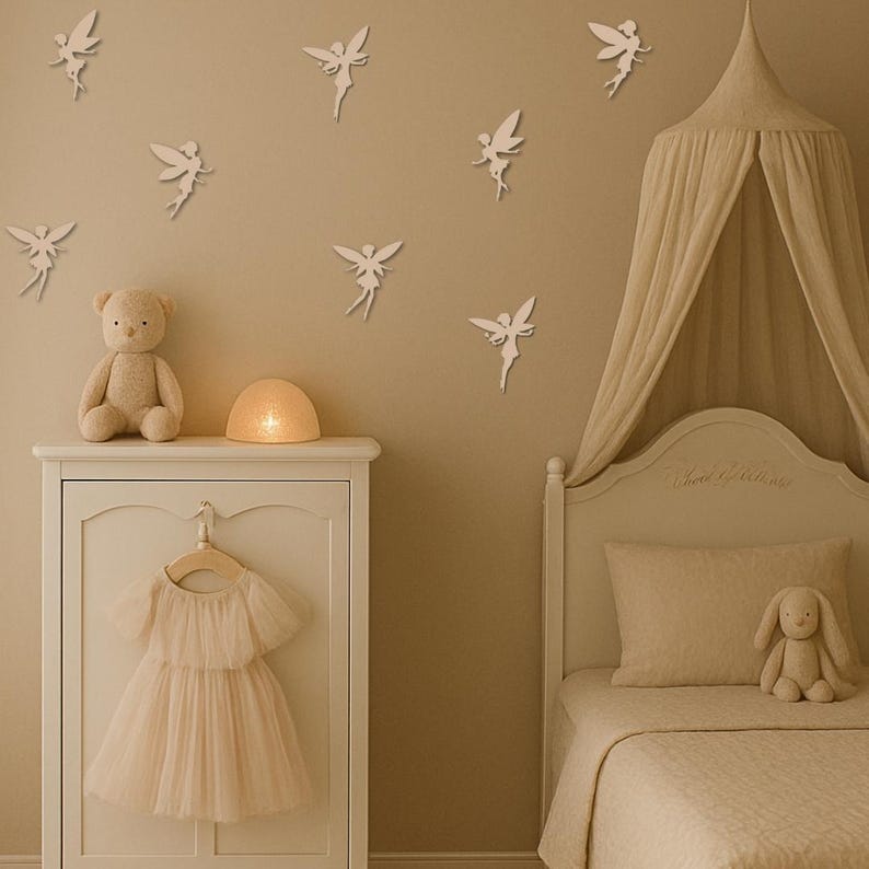 May include: A nursery scene with a white dresser, a bed with a canopy, and fairy wall decorations. A teddy bear and a bunny plush sit on the dresser. A light pink dress hangs on a wooden hanger. The room has a soft, neutral color palette.