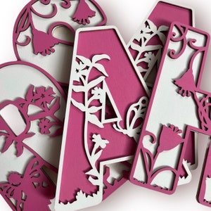 May include: Pink wooden letters with a floral design cut out. The letters are arranged in a pile, with the letter "A" on top.