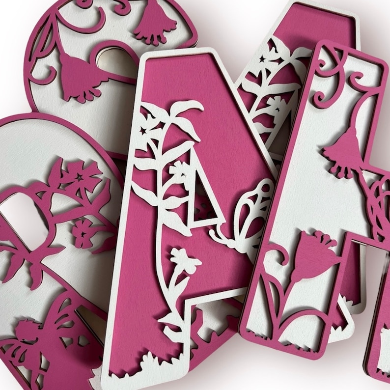 May include: Pink wooden letters with a floral design cut out. The letters are arranged in a pile, with the letter "A" on top.