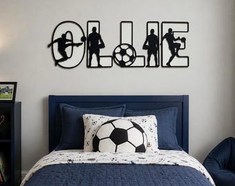 Puede incluir: Un dormitorio con una cama azul marino, una almohada con forma de balón de fútbol y una decoración de pared que deletrea "OLLIE" con siluetas de futbolistas. La ropa de cama incluye un edredón estampado y fundas de almohada a juego.
