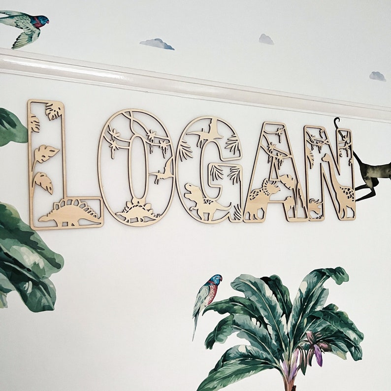 May include: Wooden wall decor with the name "LOGAN" spelled out in large letters. Each letter is decorated with a jungle theme, featuring dinosaurs, palm trees, and vines.