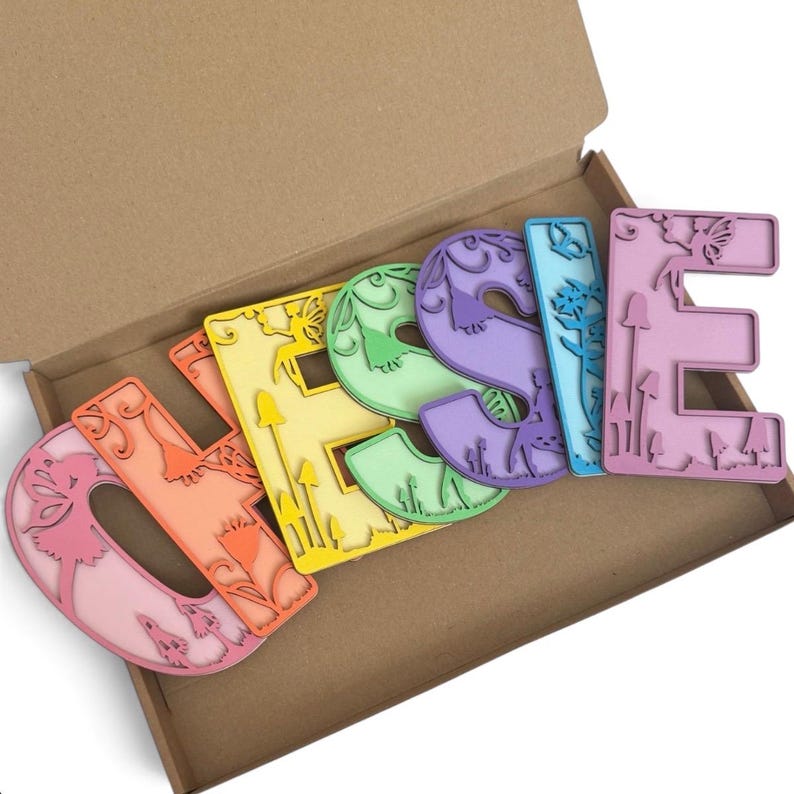 May include: A set of six colorful wooden letters spelling the name JESSIE, each featuring intricate laser-cut designs of fairies, mushrooms, and floral patterns. The letters are arranged in a cardboard box, showcasing a gradient of pastel colors.