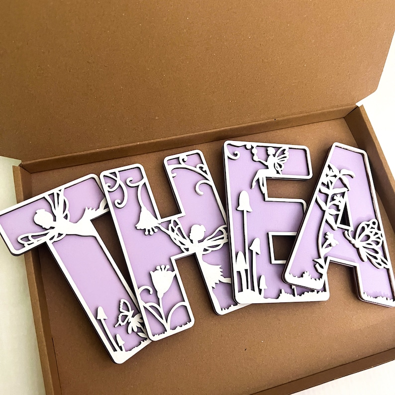 May include: Purple wooden letters spelling "THEA" with a fairy and floral design. The letters are in a box.