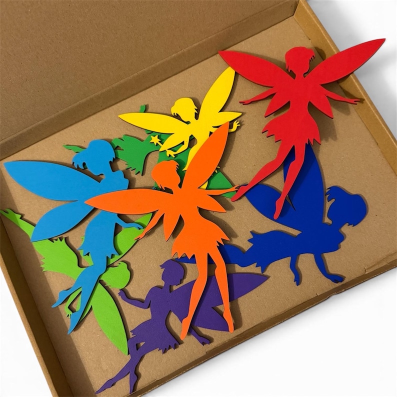 May include: A collection of colorful fairy cutouts in a cardboard box. The fairies are in various colors, including red, orange, yellow, green, blue, and purple. Each fairy has detailed wings and a silhouette design.