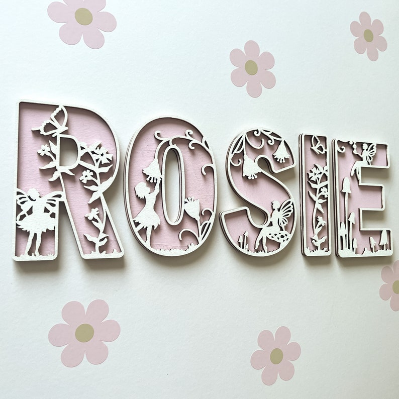 May include: Pink wooden letters spelling the name "ROSIE" with a fairy and floral design. Each letter has a cutout design of a fairy and flowers.