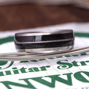 Puede incluir: Primer plano de un anillo con una banda central negra y un borde de metal plateado. El anillo está sobre una cuerda de guitarra y una superficie impresa en verde y blanco.