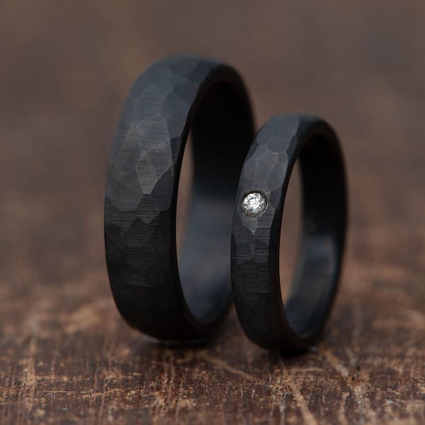 May include: Two black rings with a hammered, textured finish. The wider ring is plain, while the narrower ring features a single, small, round, clear gemstone set flush into the center of the band, creating a distinct contrast against the dark metal.