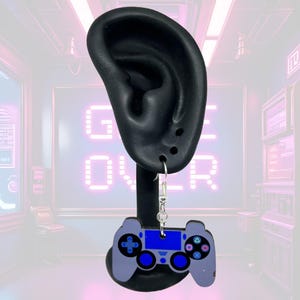 May include: A pair of dangle earrings featuring a video game controller design. The earrings are light purple with blue accents and a silver-colored hook. The controller has buttons and directional pads.