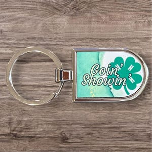 May include: Silver-toned metal keychain with a rectangular charm. The charm features a green and white design with the words "Goin' Showin'" and a four-leaf clover. The background has a watercolor effect with shades of green and gold.
