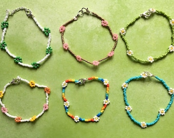 May include: Six beaded bracelets with flower charms. The bracelets are made with white, green, pink, orange, yellow, blue, and turquoise beads. The flower charms are white with yellow centers.