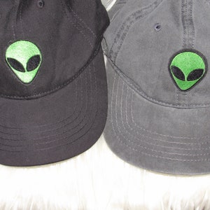 May include: Two baseball caps with an alien face embroidered on the front. The cap on the left is black and the cap on the right is gray.