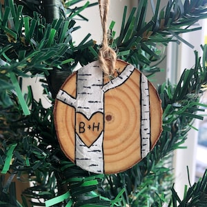 May include: A wooden ornament shaped like a birch tree with a heart carved into the trunk. The heart has the initials "B+H" written inside. The ornament is hanging from a string on a green artificial Christmas tree.