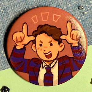May include: A round, pink button featuring a cartoon illustration of a person in a suit making a hand gesture. The person has brown hair and is smiling. The suit is blue and maroon striped. Three small, open rectangles are above the person's head.