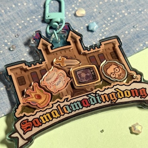 May include: A clear acrylic keychain featuring a castle design with various cartoon images and the text "Samalamadingdong". The keychain has a teal clasp and includes a crown, a sword, a TV, and a character's face.