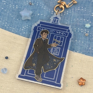 Könnte beinhalten: Ein klarer Acryl-Schlüsselanhänger mit einem blauen TARDIS-Design und einer Charakterillustration. Der Schlüsselanhänger hat einen goldfarbenen sternförmigen Anhänger und einen Metallring. Die Figur trägt einen dunklen Mantel mit Sternenmustern.