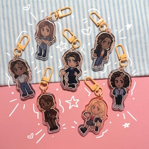 May include: A collection of seven acrylic keychains featuring cartoon illustrations of characters in various outfits. Each keychain has a gold-colored clasp. The background is pink with white star and heart doodles, and a blue and white striped pattern.