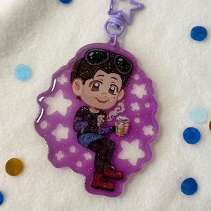 May include: Purple glitter keychain featuring a cartoon character wearing sunglasses and a black shirt, holding a cup of coffee. The character is surrounded by white stars.