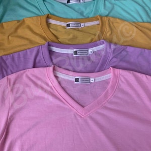 May include: A collection of V-neck t-shirts in various pastel colors: light blue, mint green, mustard yellow, lavender, and pink. Each shirt has a white tag with the brand name. The shirts are neatly arranged, showcasing their different hues.