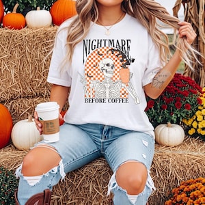 Può includere: Una t-shirt bianca a maniche corte con la grafica di uno scheletro che tiene in mano una tazza di caffè. Il design include un arco a scacchi arancione e bianco, pipistrelli neri e il testo "NIGHTMARE BEFORE COFFEE" in un font nero e audace.