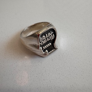May include: A silver-toned ring featuring a raised, stylized head-shaped emblem on the face. The emblem contains the embossed text "BRAIN DEAD" above a row of four small, circular dots, all set against a recessed, darkened background.